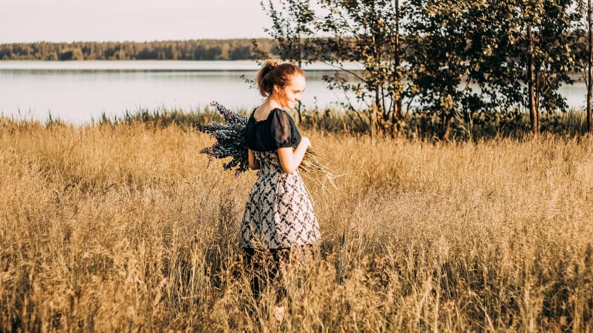 A young woman stands in a sunlit field by a lake, holding lavender flowers and smiling.
